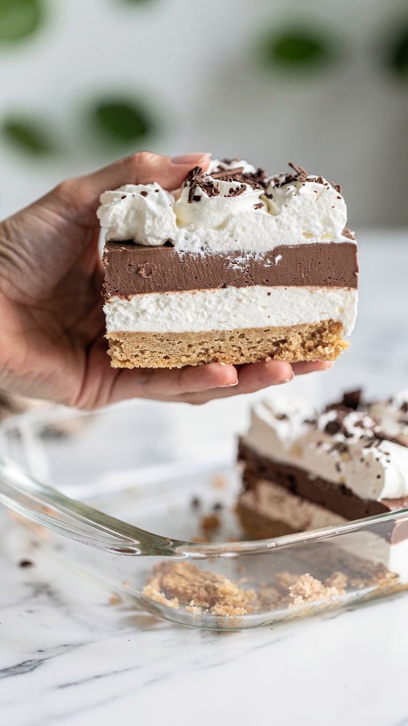 The image shows a close-up of a layered dessert held by a woman's hand above a clear glass dish. The dessert has four layers: the bottom layer is a light brown crumbly base, the second layer is a thick white creamy filling, the third layer is a smooth dark brown chocolate layer, and the top layer is fluffy white whipped cream sprinkled with small chocolate shavings. The clear dish below contains the rest of the dessert with a portion missing where the dessert is held. The background features a white marbled surface and green blurred shapes. Photo taken with an iphone --ar 4:5 --v 7