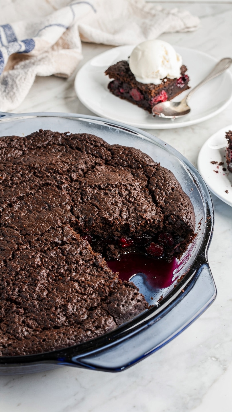 The image shows one piece of dessert on a white plate with a white marbled surface underneath. The dessert has two main layers: the top layer is a dark brown, rough, and crumbly cake-like texture, while the bottom layer is a thick, shiny red berry sauce with visible whole berries, creating a glossy and juicy look. A silver fork is placed on the right side of the plate, with a woman’s hand holding it gently. The plate looks slightly shiny from the light. photo taken with an iphone --ar 4:5 --v 7