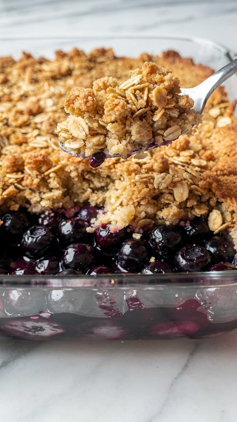 This image shows a round clear glass dish filled with a two-layer dessert. The bottom layer is a deep purple blueberry fruit filling, thick and glossy with whole blueberries visible. The top layer is a golden-brown crumb topping made of oats and crumbly bits unevenly spread across the fruit. A metal spoon rests on the left side, slightly digging into the dish, with some blueberry filling on it. The dish is placed on a white marbled surface. photo taken with an iphone --ar 4:5 --v 7